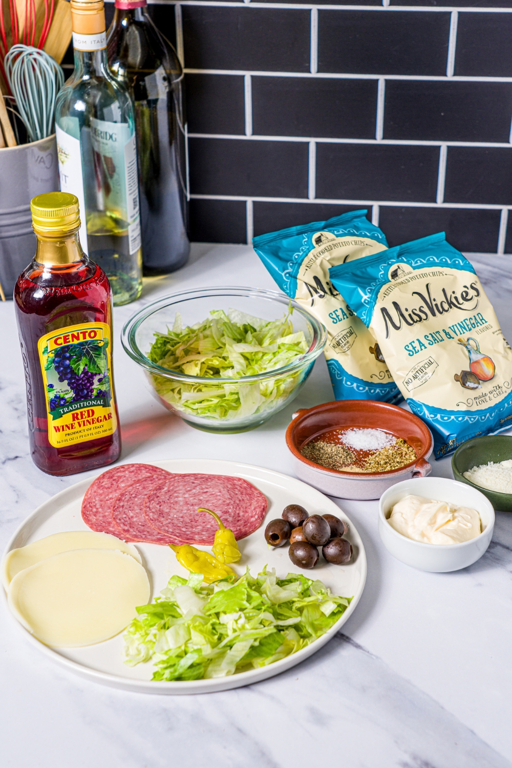 A white plate of salami, provolone, lettuce, and black olives. Behind that is a bowl of shredded lettuce, two salt vinegar, chip bags, a small bowl of Italian seasonings, a small bowl of mayo, and a bottle of vinegar.