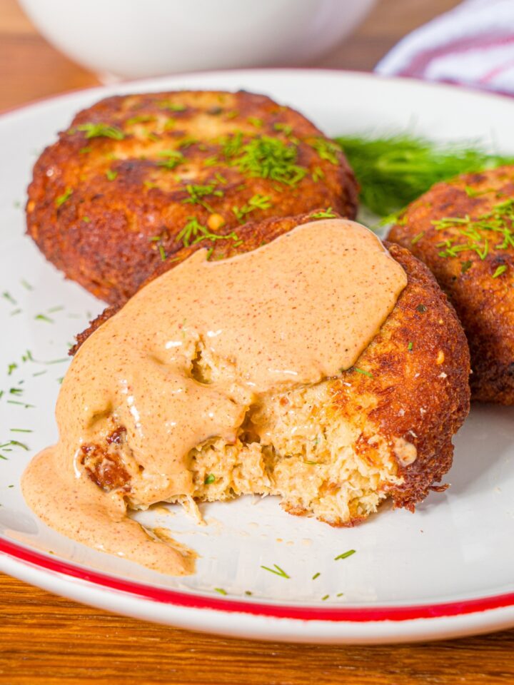 A Maryland style crab cake with remoulade sauce on top of it and a piece taken out from the front of it on a white plate with two more crab cakes.