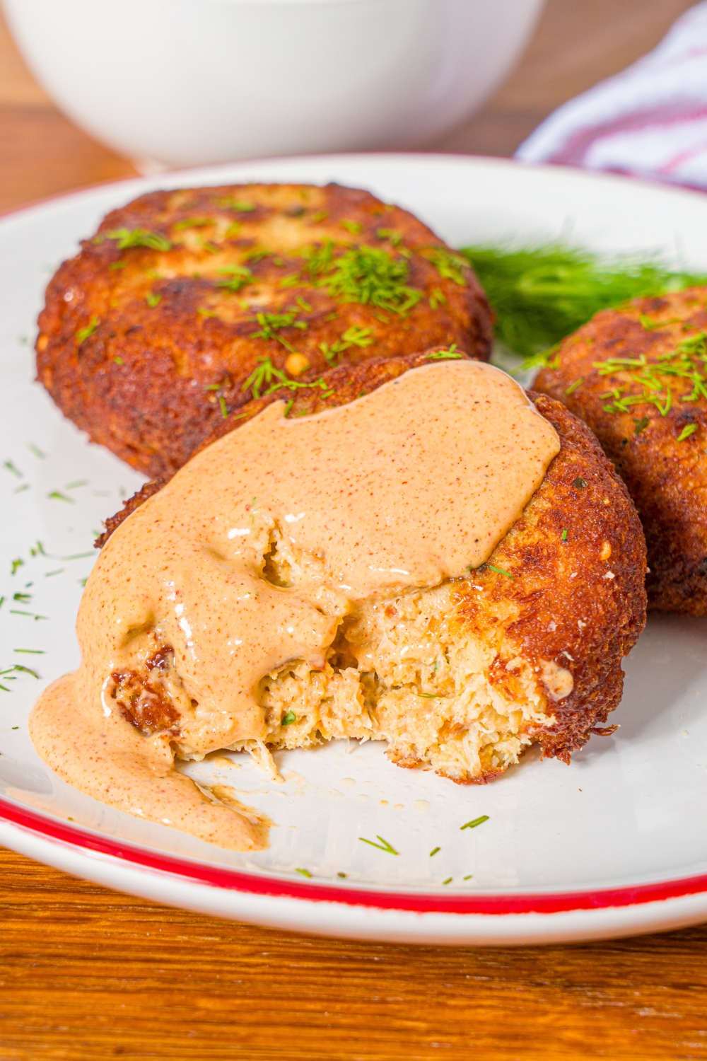 A Maryland style crab cake with remoulade sauce on top of it and a piece taken out from the front of it on a white plate with two more crab cakes.