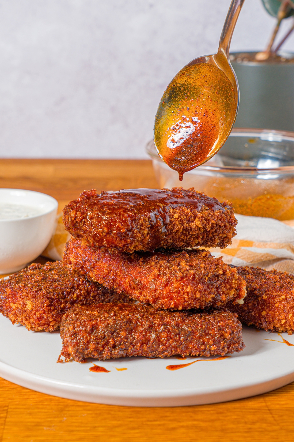 A spoon drizzling Nashville hot sauce on top of a flat mozzarella stick that is stacked on top of four more mozzarella sticks.