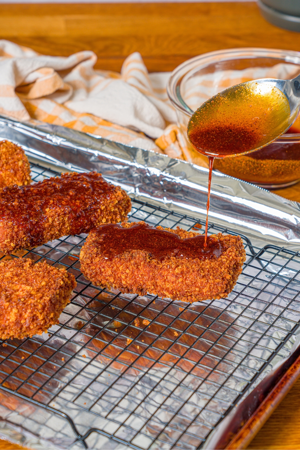 A spoon pouring Nashville hot sauce on top of a flat mozzarella stick on a wire rack with more Nashville hot mozzarella sticks