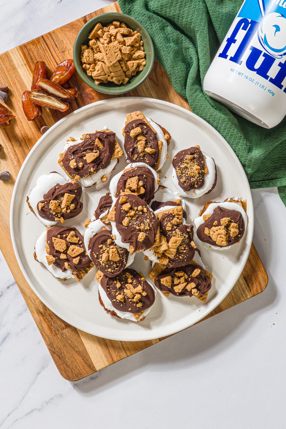 A bunch of no bake smashed s'mores that are scattered on a white plate with a few on top of one another. This white plate is on a wooden cutting board that has a bowl of crushed graham crackers and a few dates on it.