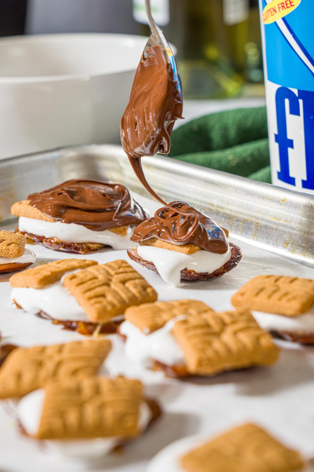 A spoon drizzling melted chocolate on top of a graham cracker that is on marshmallow fluff on a smashed date.