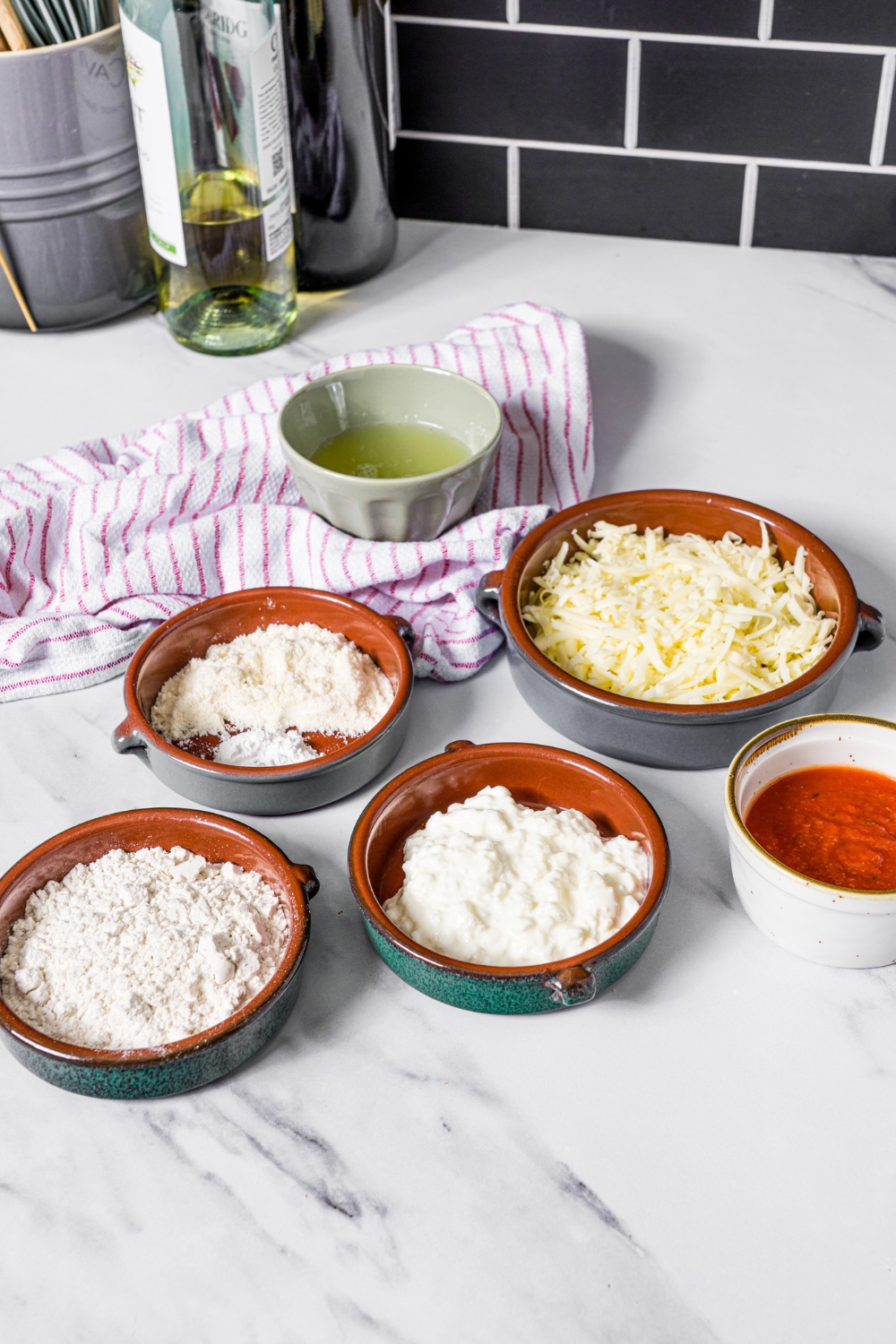 A bowl of shredded mozzarella cheese, a bowl of gluten-free flour, a bowl of coconut flour, a bowl of cottage cheese, and a bowl of marinara sauce on a white counter.