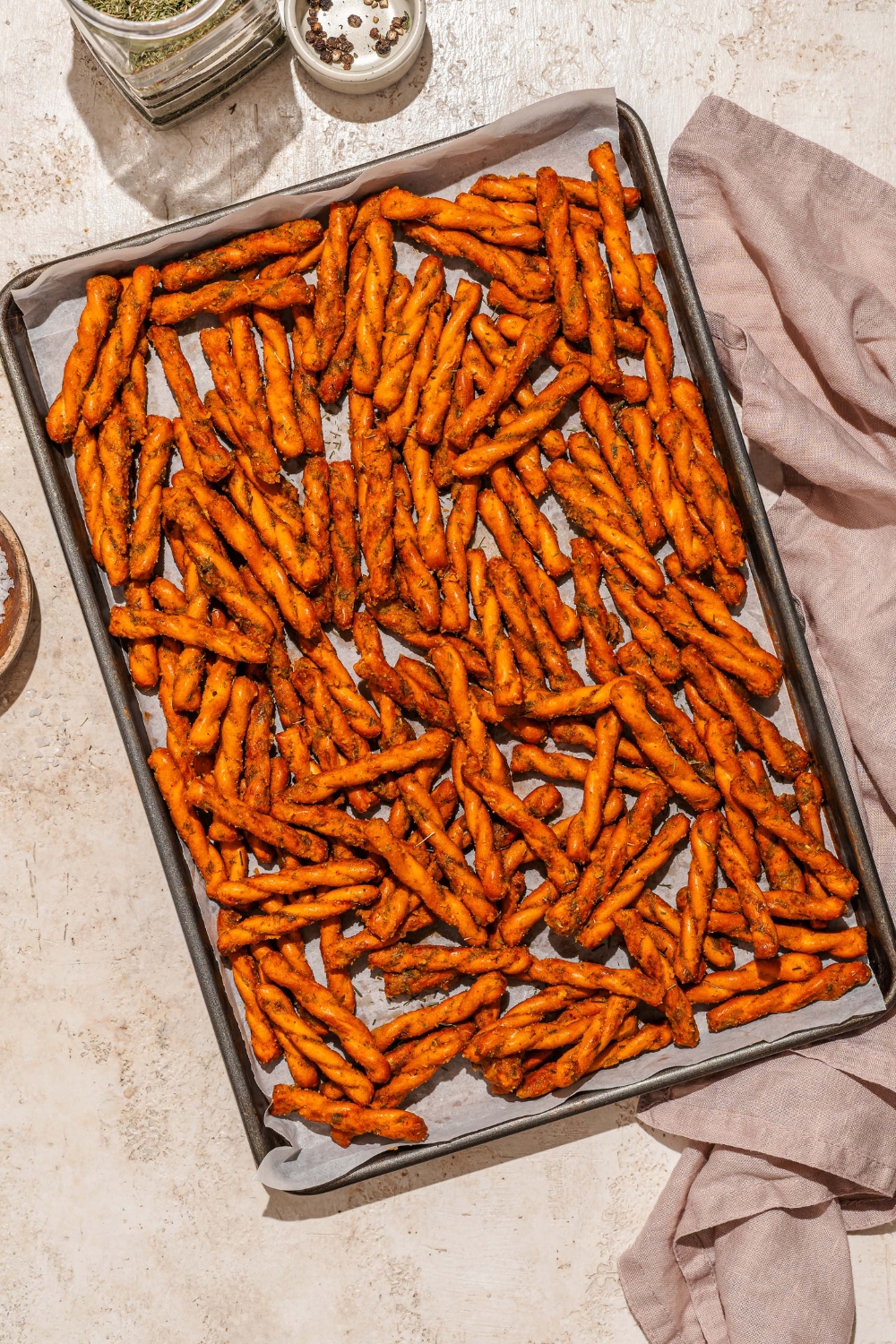 A baking sheet lined with parchment paper with seasoned pretzel braids. The sheet is on a tan counter with a white cloth napkin.