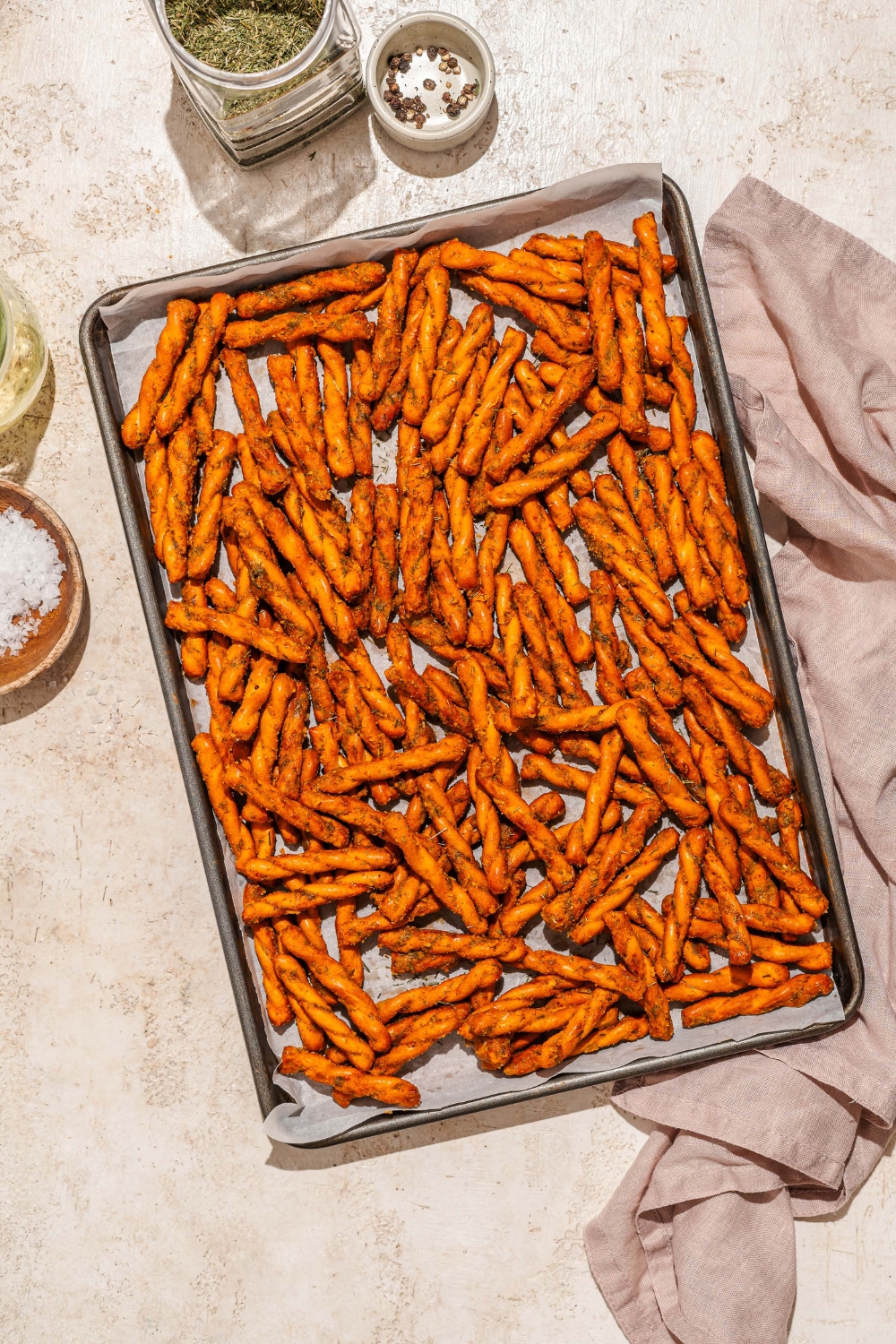 A baking sheet lined with parchment paper with seasoned pretzel braids. The sheet is on a tan counter with a white cloth napkin.