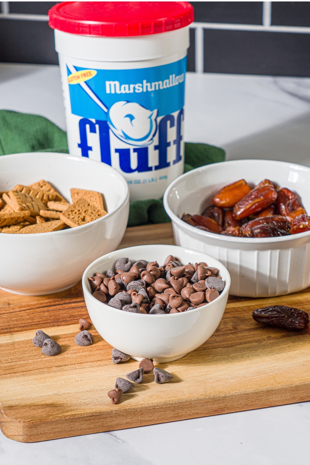 A tub of marshmallow fluff, a white bowl with broken up graham crackers, a bowl of chocolate chips, and a bowl of dates all on a wooden cutting board.