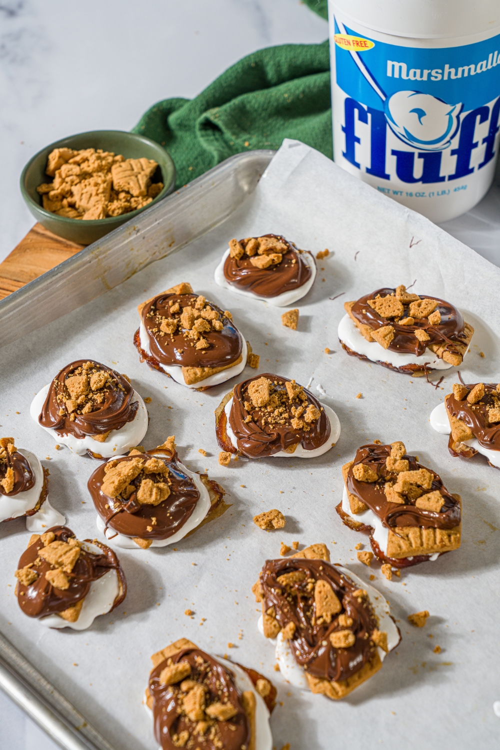 Crushed graham crackers on top of melted chocolate on marshmallow fluff on a smashed date. There are a few rows of these on a sheet of parchment paper on a baking tray.