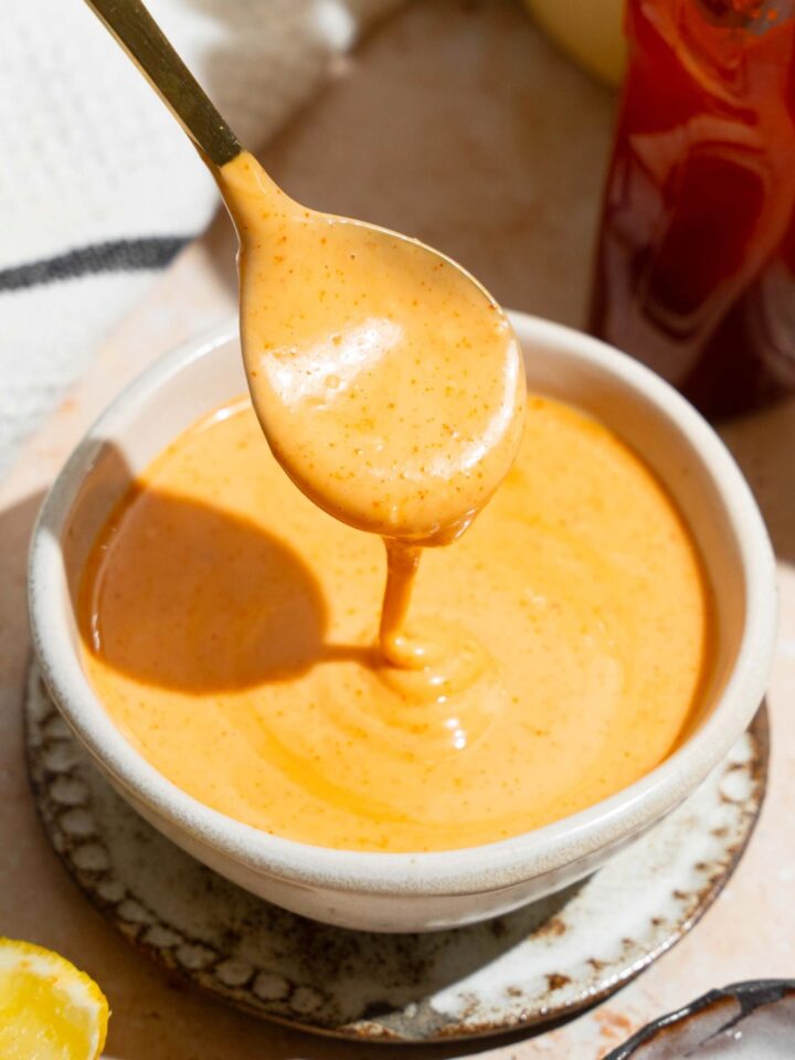 A spoon with honey sriracha sauce on it drizzling the sauce into a white bowl that is filled with the sauce.