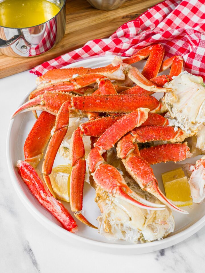 A bunch of steamed crab legs on a white plate with lemon wedges. Behind the plate is a pot of melted butter.