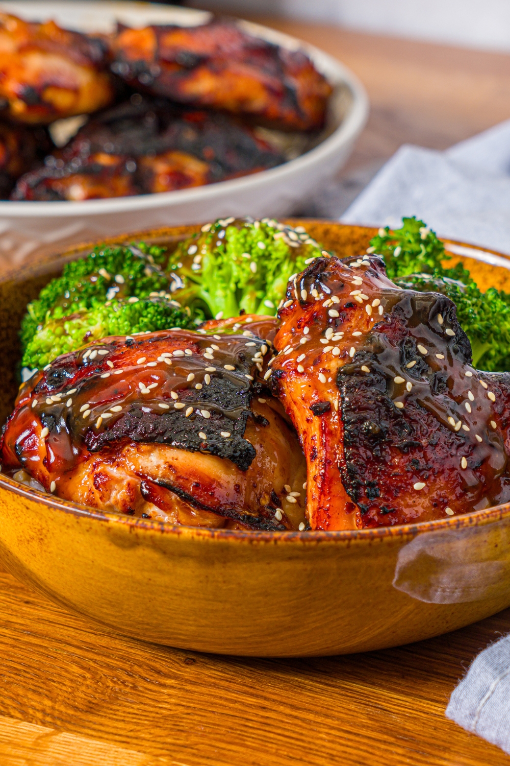 A bowl with chicken teriyaki served with broccoli and rice and garnished with sesame seeds. The bowl is on a wooden counter with a bowl of cooked chicken.