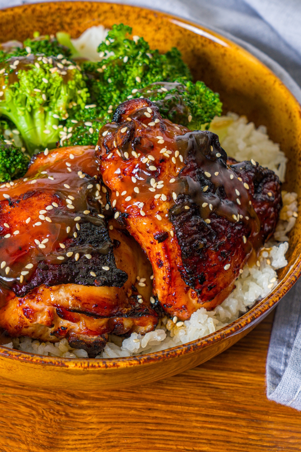 A bowl with chicken teriyaki served with broccoli and rice and garnished with sesame seeds. The bowl is on a wooden counter with a blue cloth napkin.