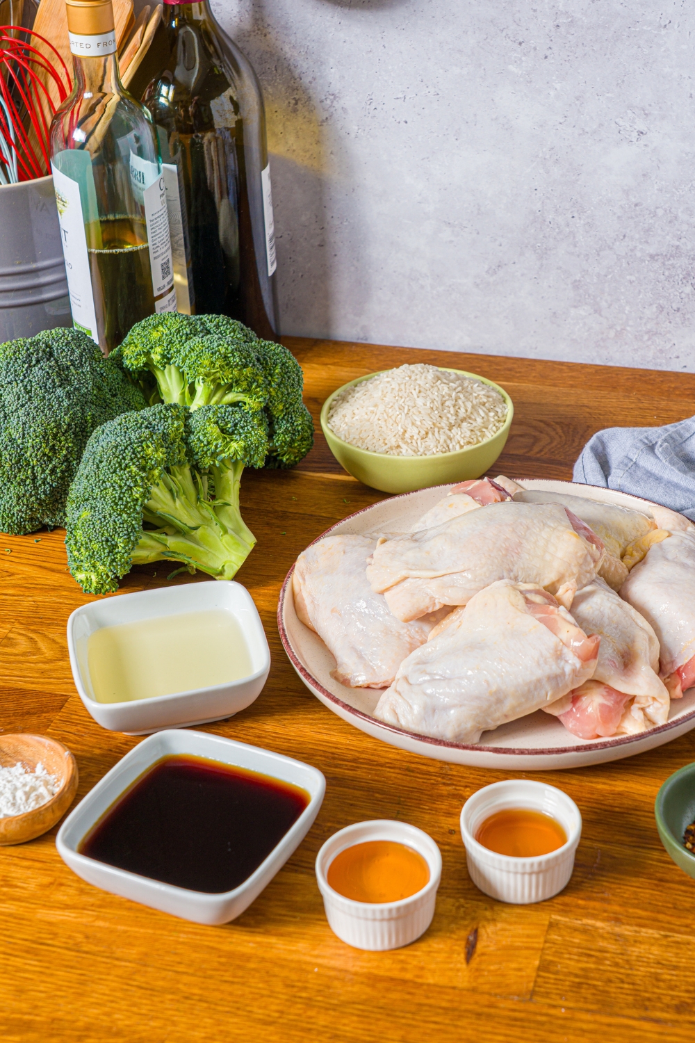 A wooden counter with several bowls containing ingredients to make chicken teriyaki bowls including chicken, rice, broccoli, garlic, oil, honey, sesame oil, and soy sauce.