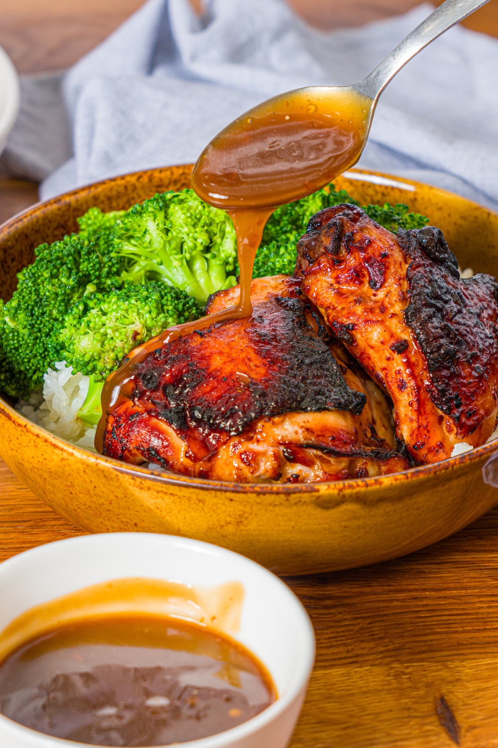 A bowl with chicken teriyaki served with broccoli and rice. A spoon is pouring teriyaki sauce over the chicken. The bowl is on a wooden counter with a blue cloth napkin and small bowl of teriyaki sauce.