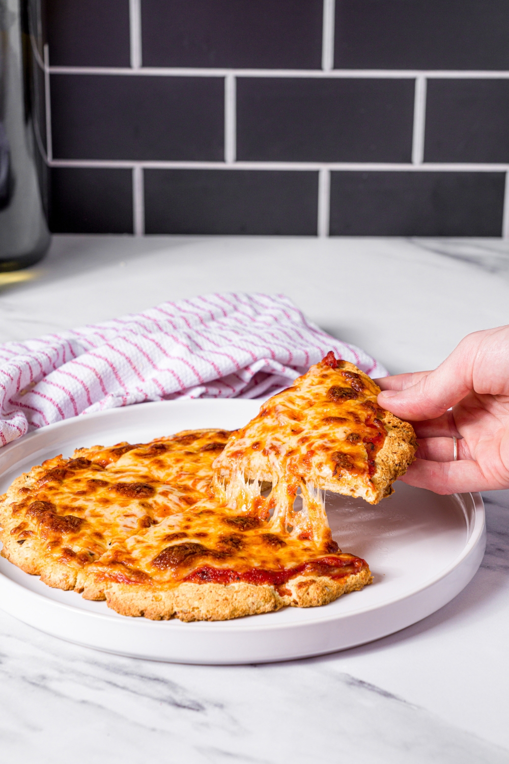 A hand pulling a slice of protein pizza away from the rest of the pizza on a white plate.
