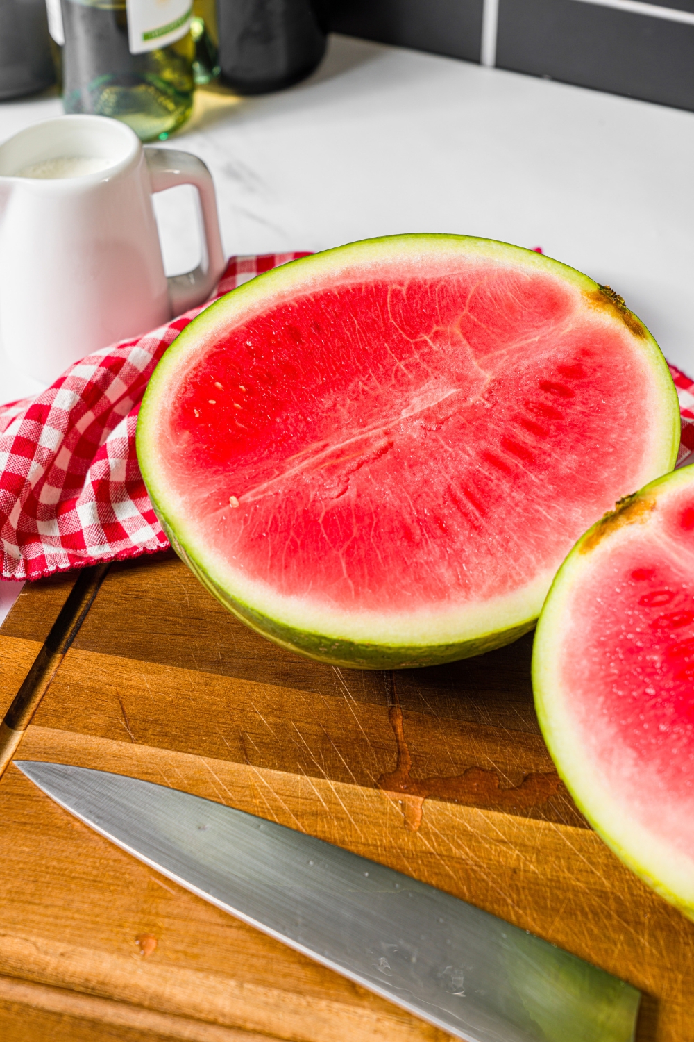 Half of a watermelon on a wooden cutting board with a pitched of heavy cream behind it.