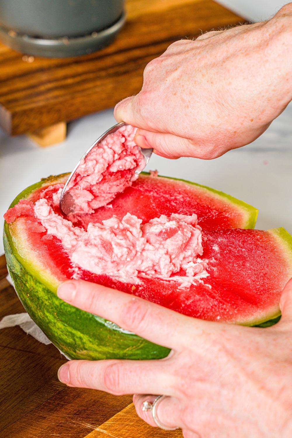A hand holding a spoon, scraping and mixing heavy cream in half of a frozen watermelon.