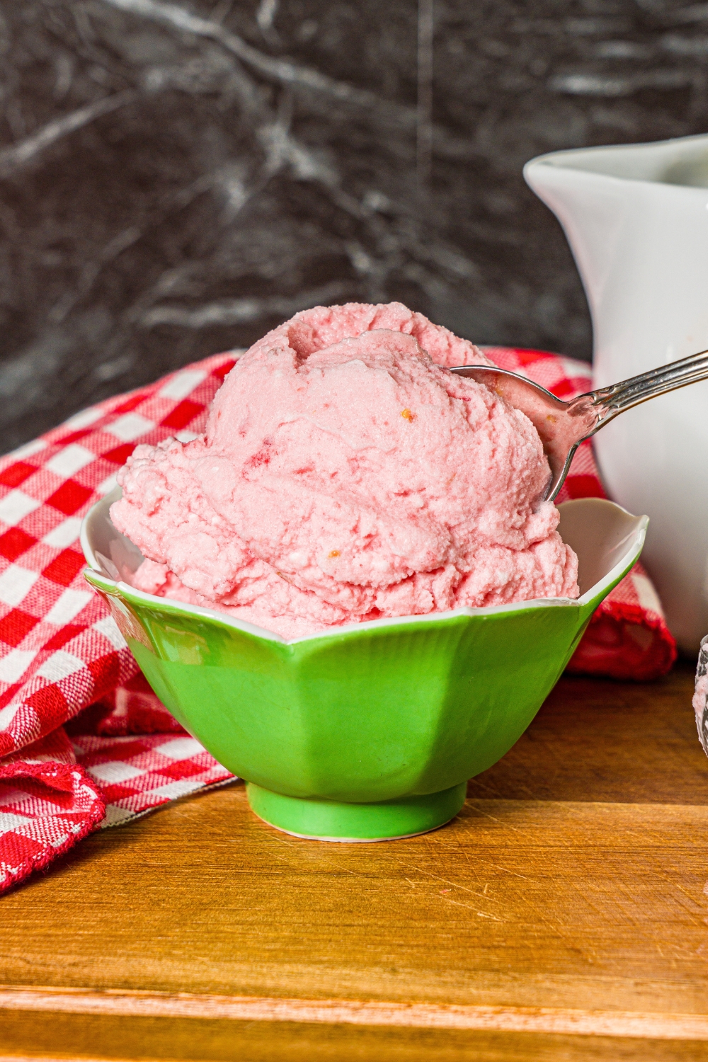 A spoon submerged in a scoop of watermelon ice cream that is in a green bowl on a wooden cutting board.