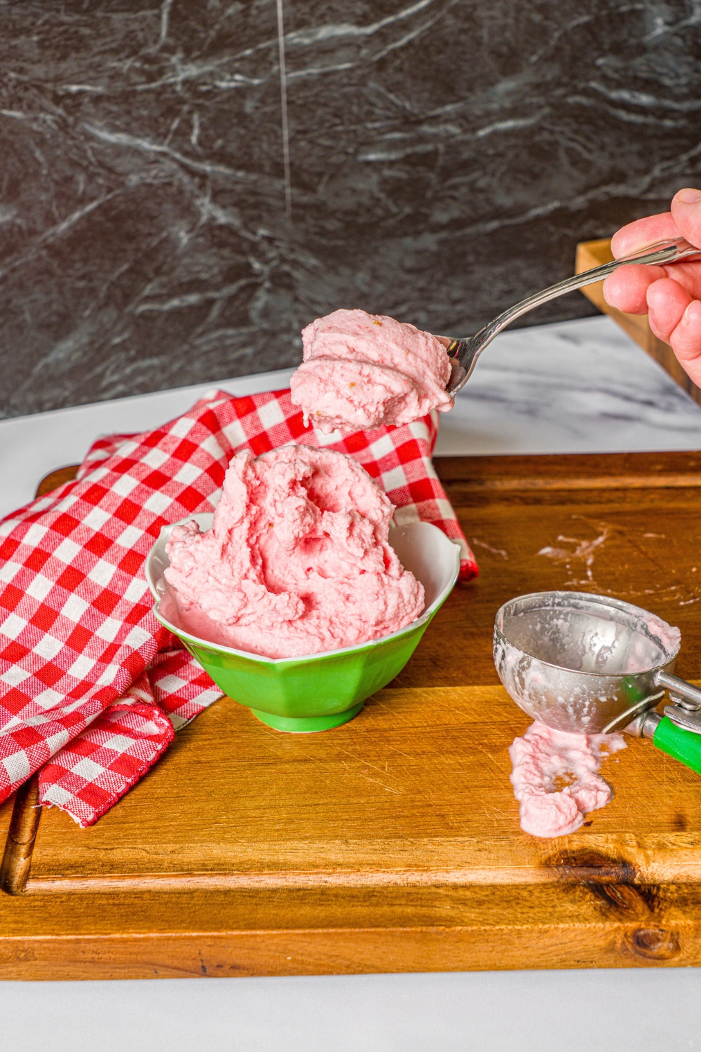 A hand holding a spoon with watermelon ice cream on it above a green bowl that is filled with the ice cream.