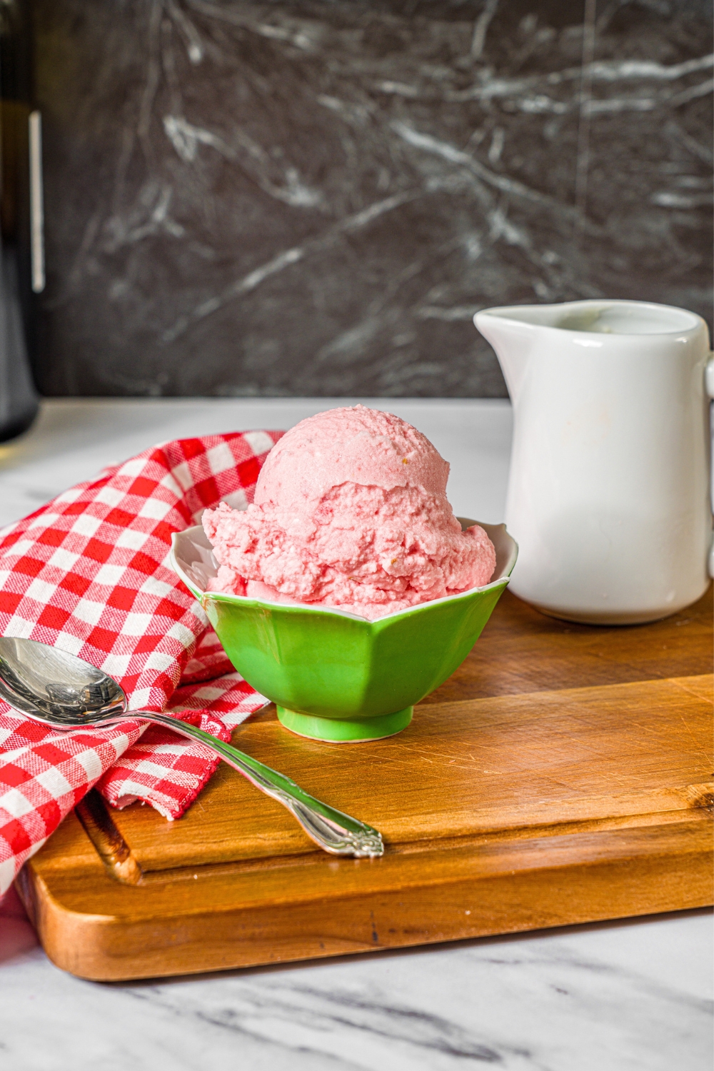 A green bowl that is filled with a scoop of watermelon ice cream on a wooden cutting board with a spoon and a white pitcher.