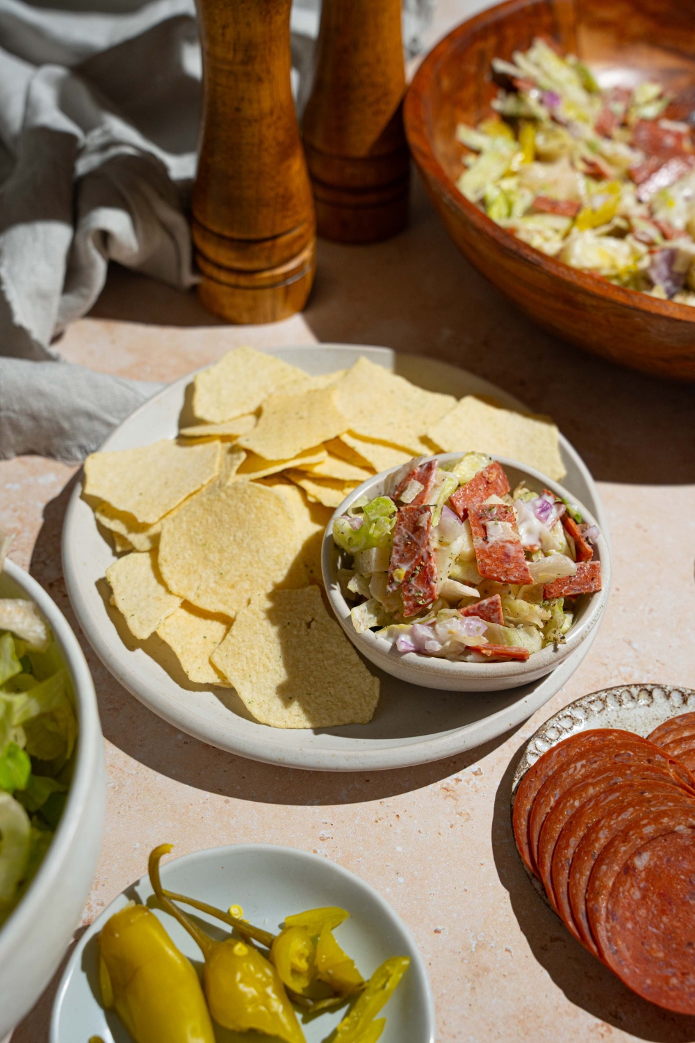A plate of chips with a small bowl of Italian grinder dip including chopped salami, pepperoni, cheese, banana peppers, and more. The plate is on a tan counter with plates of ingredients and bowl of Italian grinder dip.