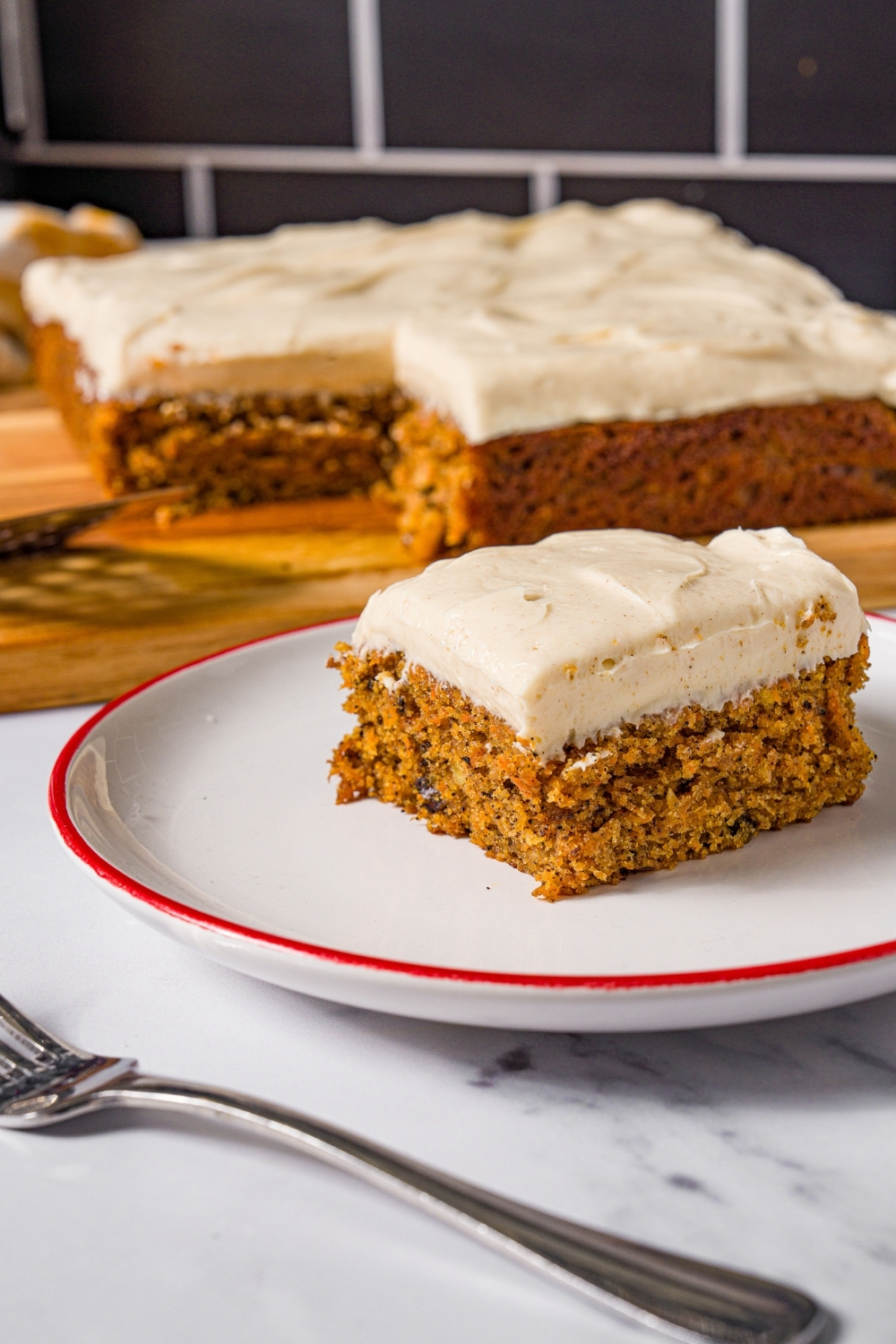 A white plate with a slice of brown butter carrot cake with cream cheese frosting. The plate is on a marble counter with a fork and a wooden board with carrot cake.