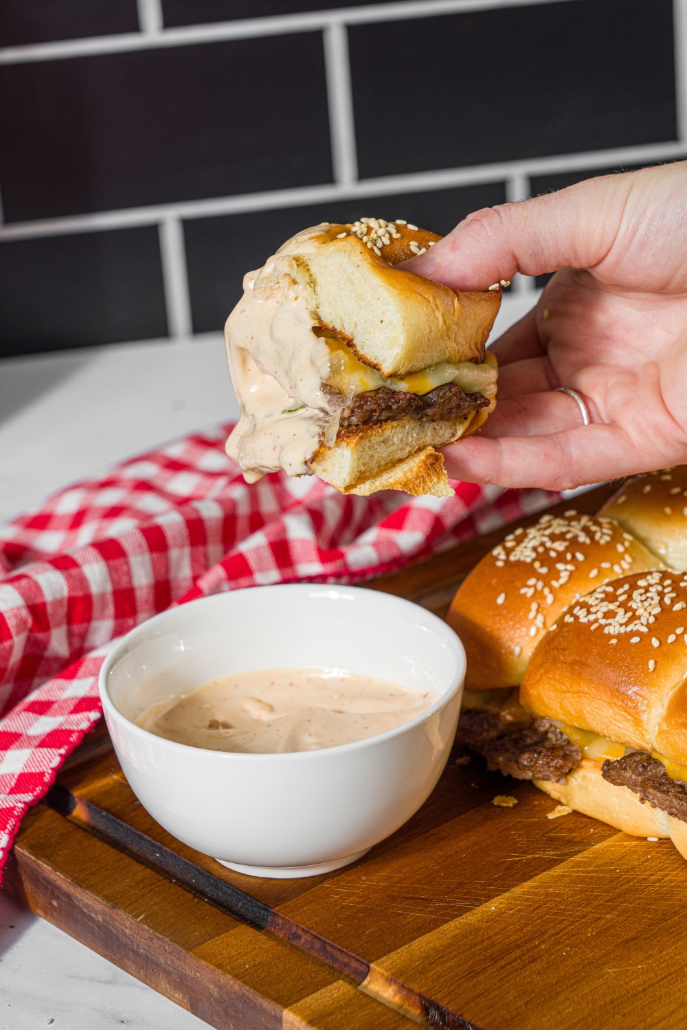 A hand holding a cheeseburger slider dipped in burger sauce over a bowl of sauce. The bowl is on a wooden board with sliders and a red checkered napkin.