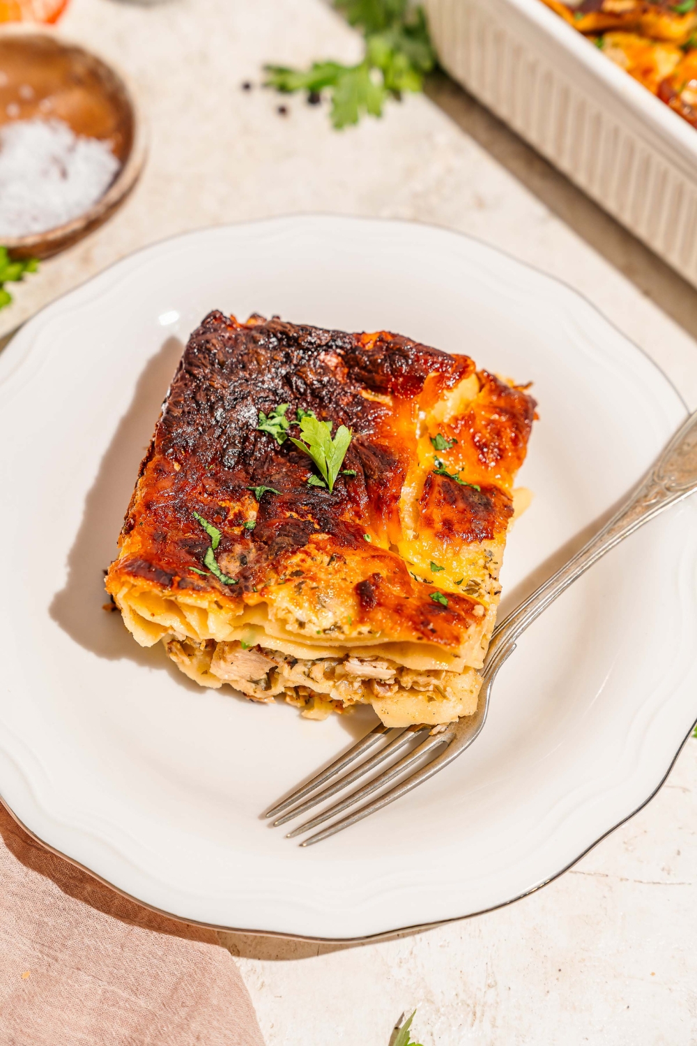 A white plate with a slice of chicken alfredo lasagna garnished with fresh parsley. There is a fork on the plate. The plate is on a tan counter with a tray of lasagna.