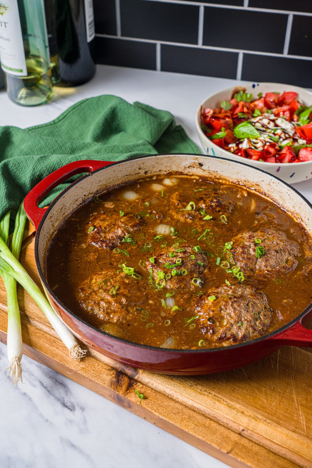A cast iron skillet with hamburger patties in onion gravy garnished with sliced green onion. The skillet is on a wooden board with a green cloth napkin.
