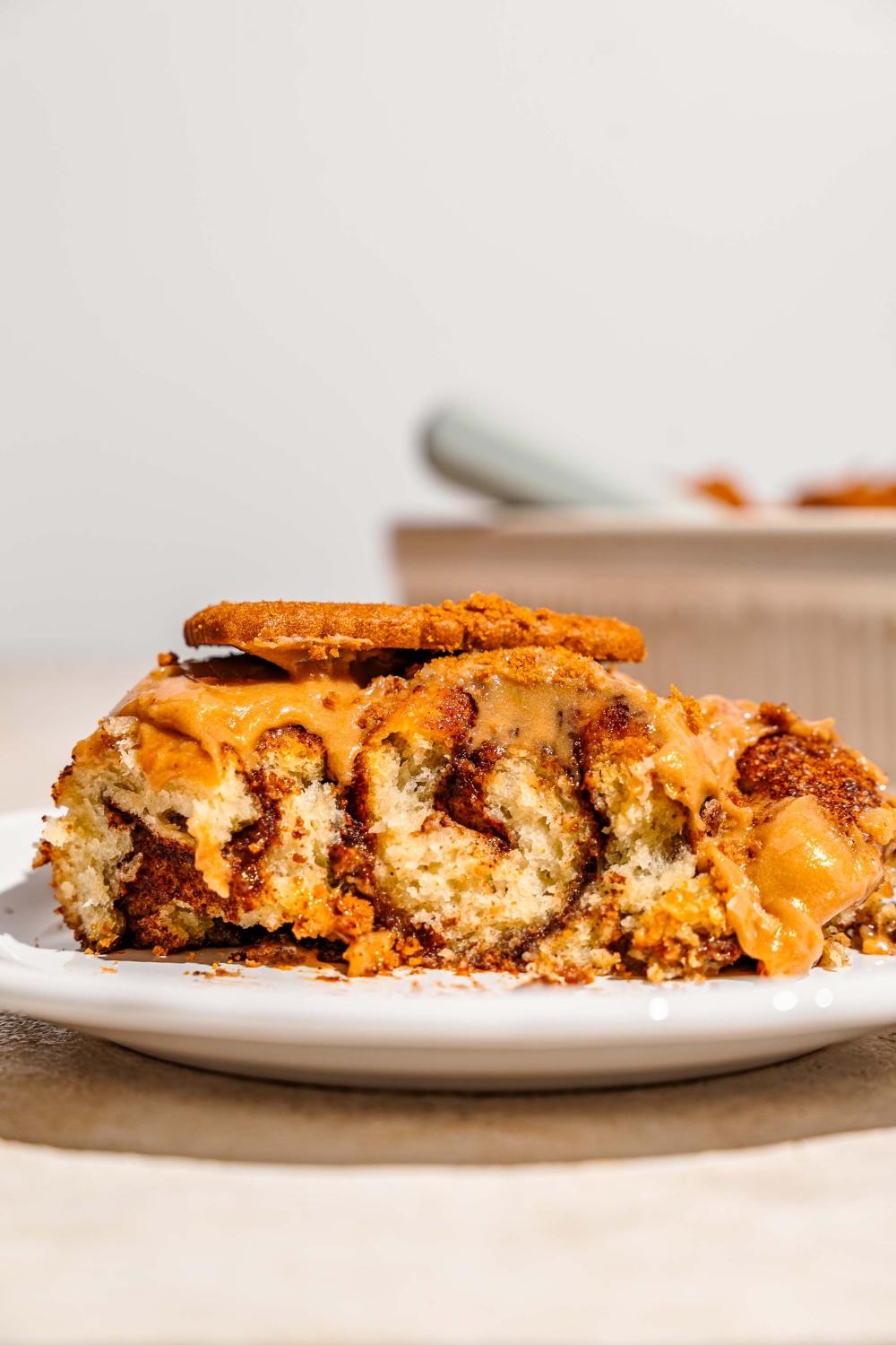 A white plate with a Biscoff cinnamon roll topped with Biscoff frosting and a whole Biscoff cookie. The plate is on a tan counter with a baking pan of cinnamon rolls.