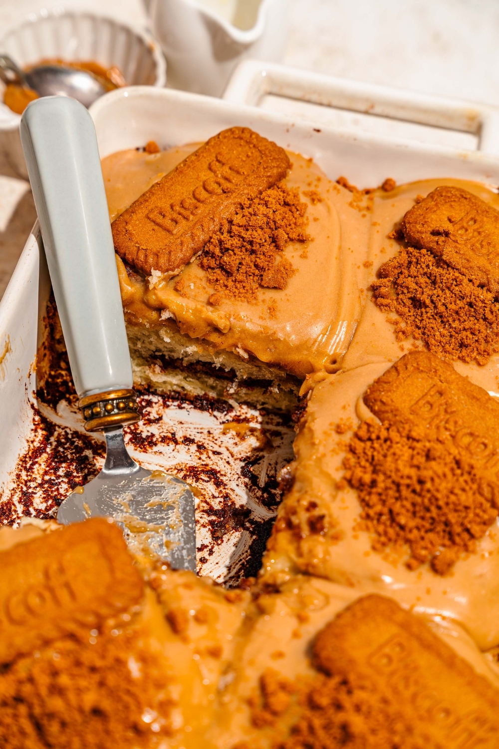A baking pan with biscoff cinnamon rolls topped with cream cheese biscoff frosting, crushed Biscoff cookies, and a whole Biscoff cookie. A cake server has taken a roll from the dish.