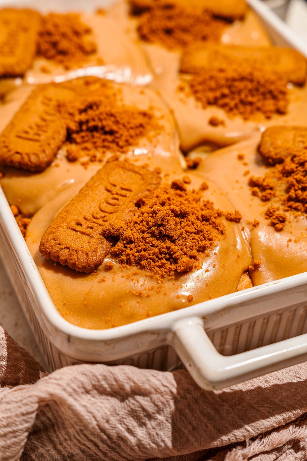 A baking pan with biscoff cinnamon rolls topped with cream cheese biscoff frosting, crushed Biscoff cookies, and a whole Biscoff cookie. The pan is on a tan counter with a brown cloth napkin.