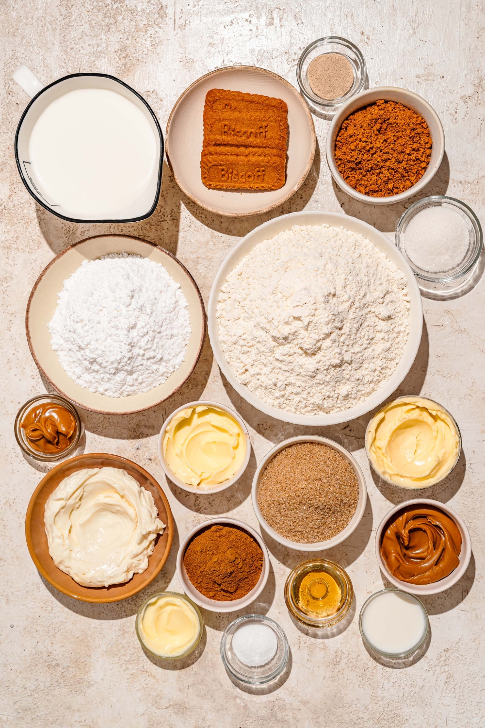 A tan counter with several bowls containing ingredients to make Biscoff cinnamon rolls including Biscoff cookies, cream cheese, cinnamon, flour, yeast, milk, butter, sugar, Biscoff cookie butter, and more.