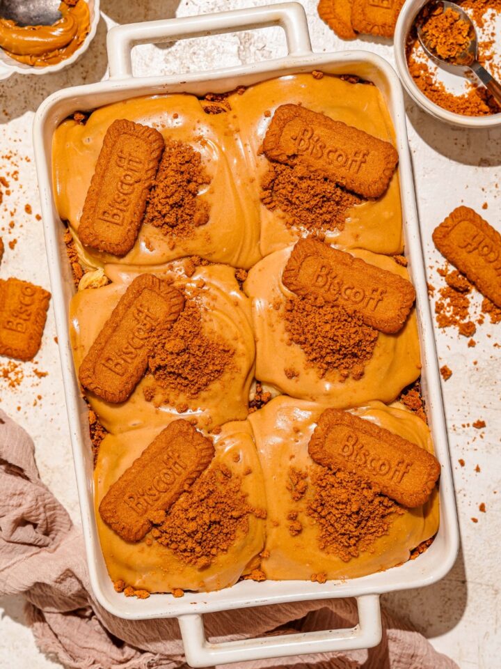 A baking pan with biscoff cinnamon rolls topped with cream cheese biscoff frosting, crushed Biscoff cookies, and a whole Biscoff cookie. The pan is on a tan counter with a brown cloth napkin and bowls of toppings.