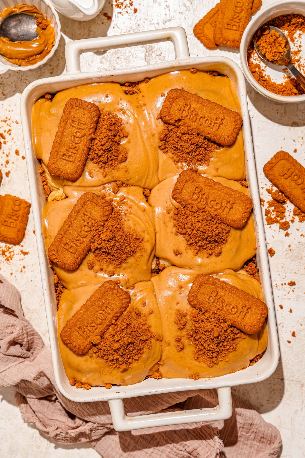 A baking pan with biscoff cinnamon rolls topped with cream cheese biscoff frosting, crushed Biscoff cookies, and a whole Biscoff cookie. The pan is on a tan counter with a brown cloth napkin and bowls of toppings.