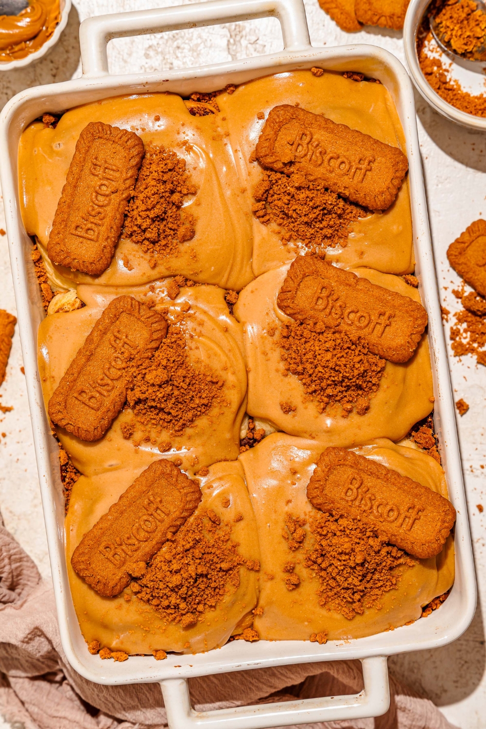 A baking pan with biscoff cinnamon rolls topped with cream cheese biscoff frosting, crushed Biscoff cookies, and a whole Biscoff cookie. The pan is on a tan counter with a brown cloth napkin.