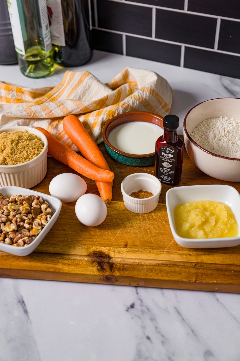 A wooden board with ingredients to make brown butter carrot cake including butter, carrots, eggs, brown sugar, walnuts, flour, vanilla, cream cheese, cinnamon, apple sauce, and more.