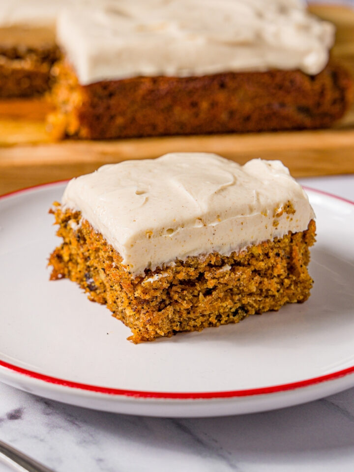A white plate with a slice of brown butter carrot cake with cream cheese frosting. The plate is on a marble counter with a fork and a wooden board with carrot cake.