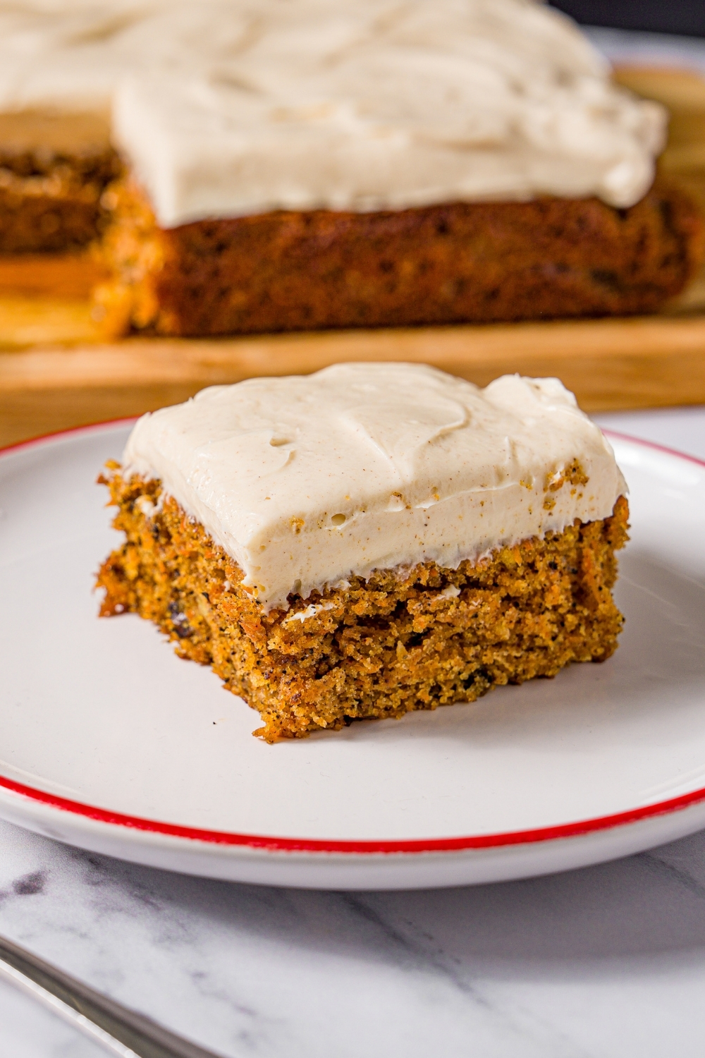 A white plate with a slice of brown butter carrot cake with cream cheese frosting. The plate is on a marble counter with a fork and a wooden board with carrot cake.