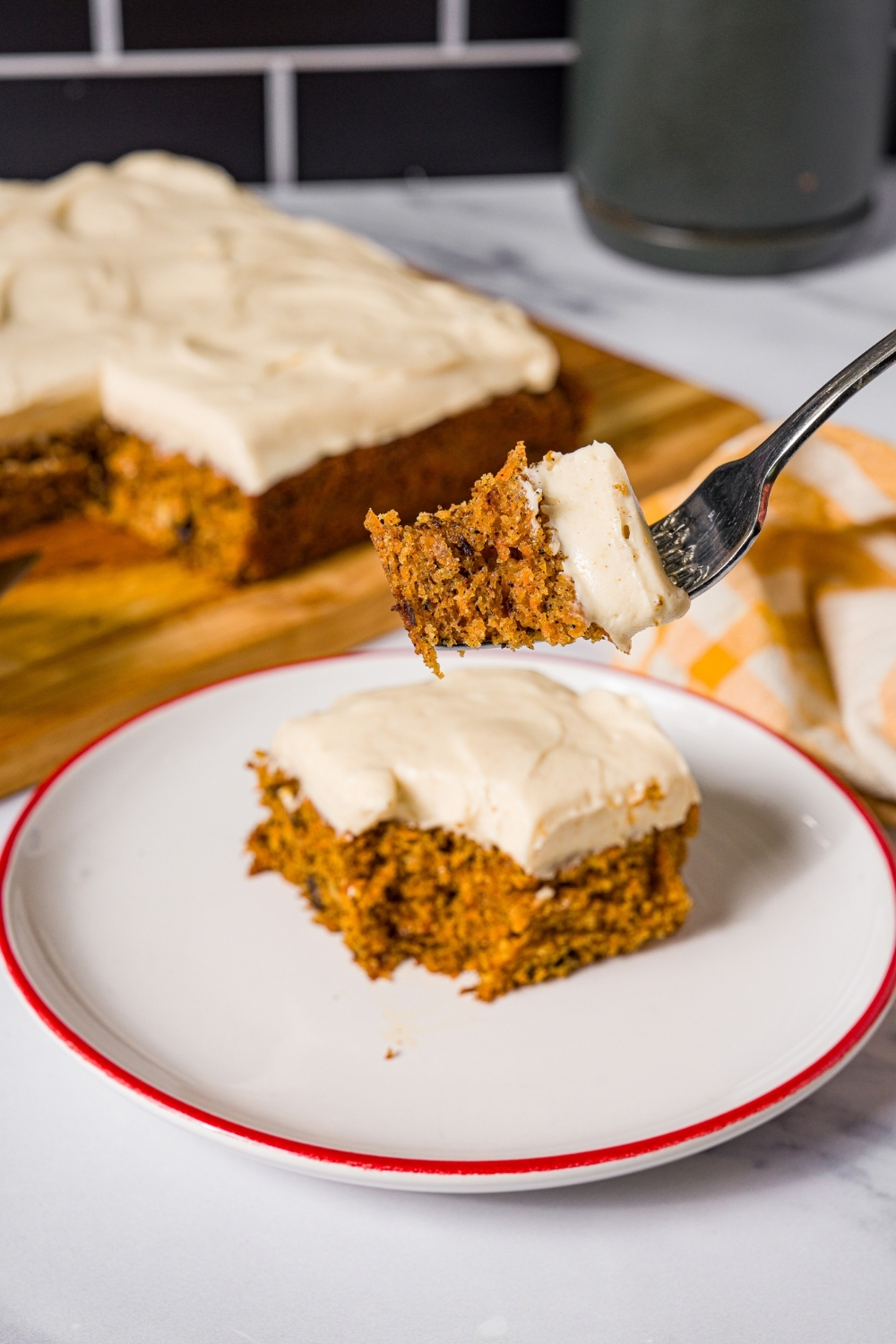 A white plate with a slice of brown butter carrot cake with cream cheese frosting. A fork is taking a bite from the cake. The plate is on a marble counter with a wooden board with a carrot cake.