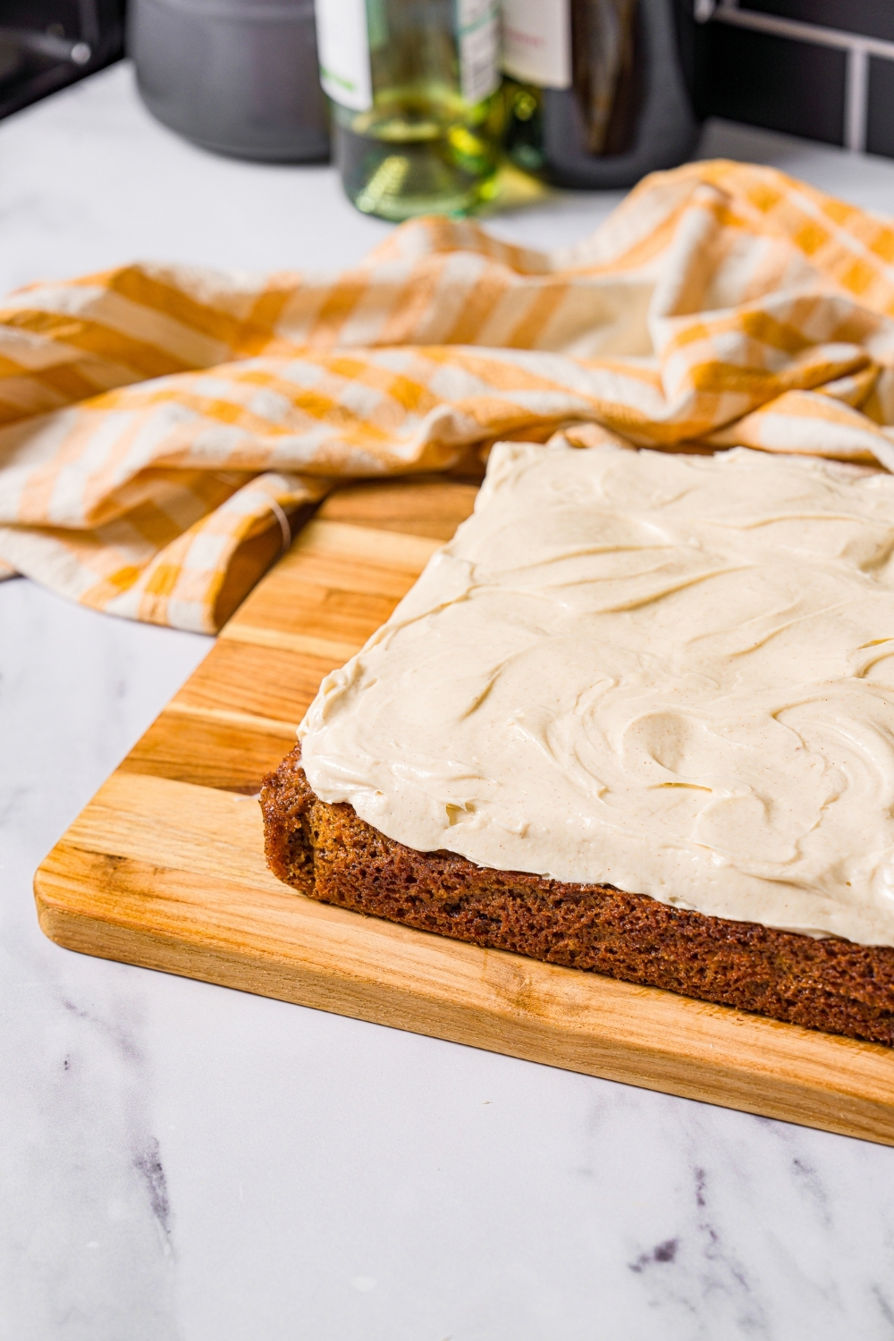 A wooden board with a whole carrot cake with cream cheese frosting. The board is on a marble counter with a white striped napkin.
