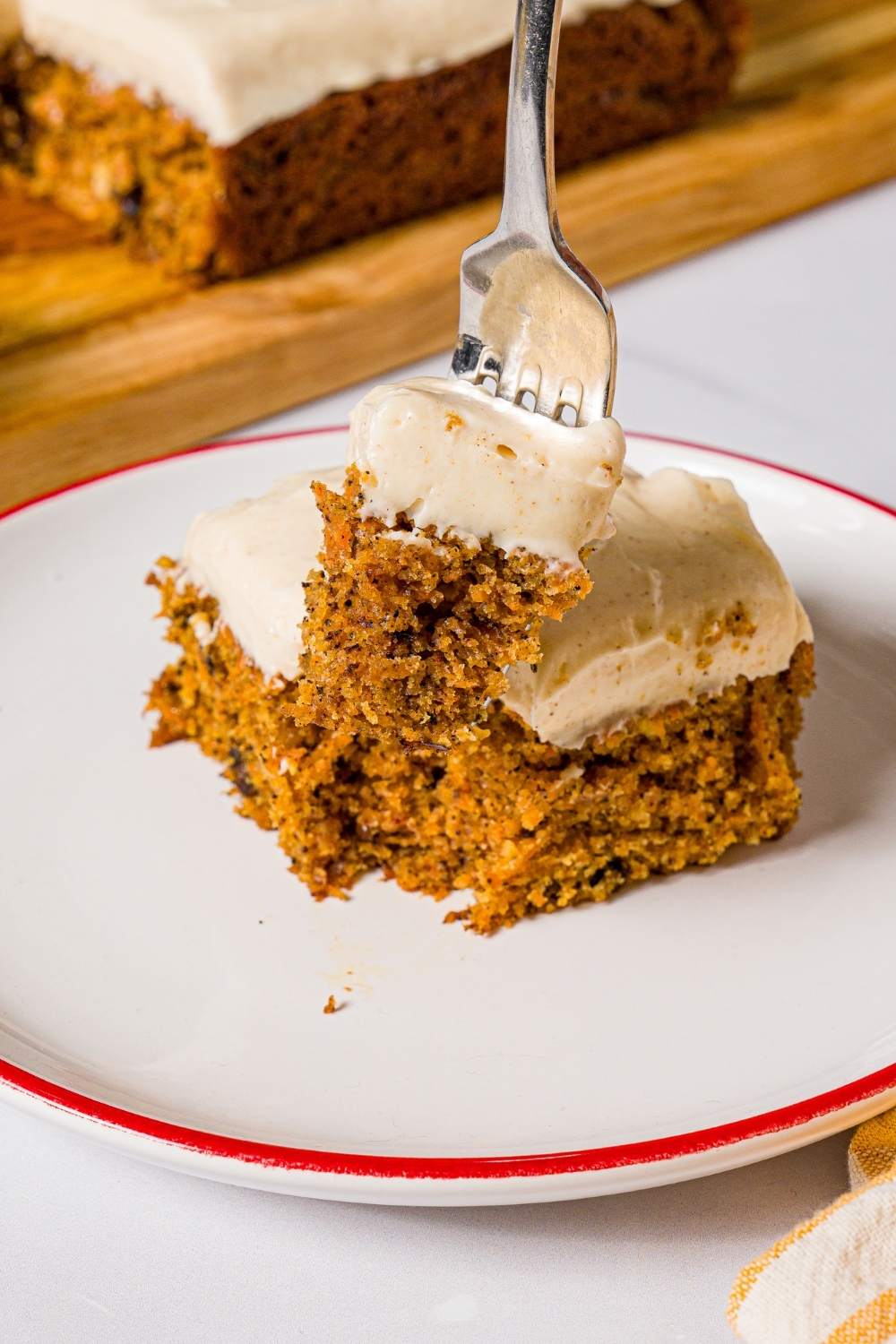 A white plate with a slice of carrot cake with cream cheese frosting. A fork is taking a bite from the cake. The plate is on a marble counter.