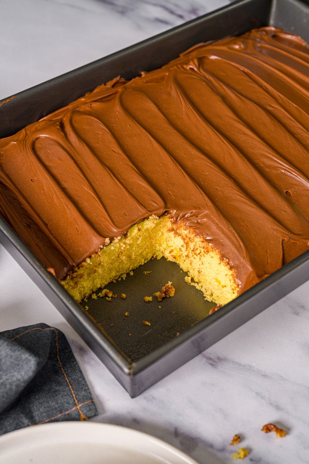 A baking pan with yellow cake with chocolate frosting. A slice is removed from the cake. The pan is on a marble counter.