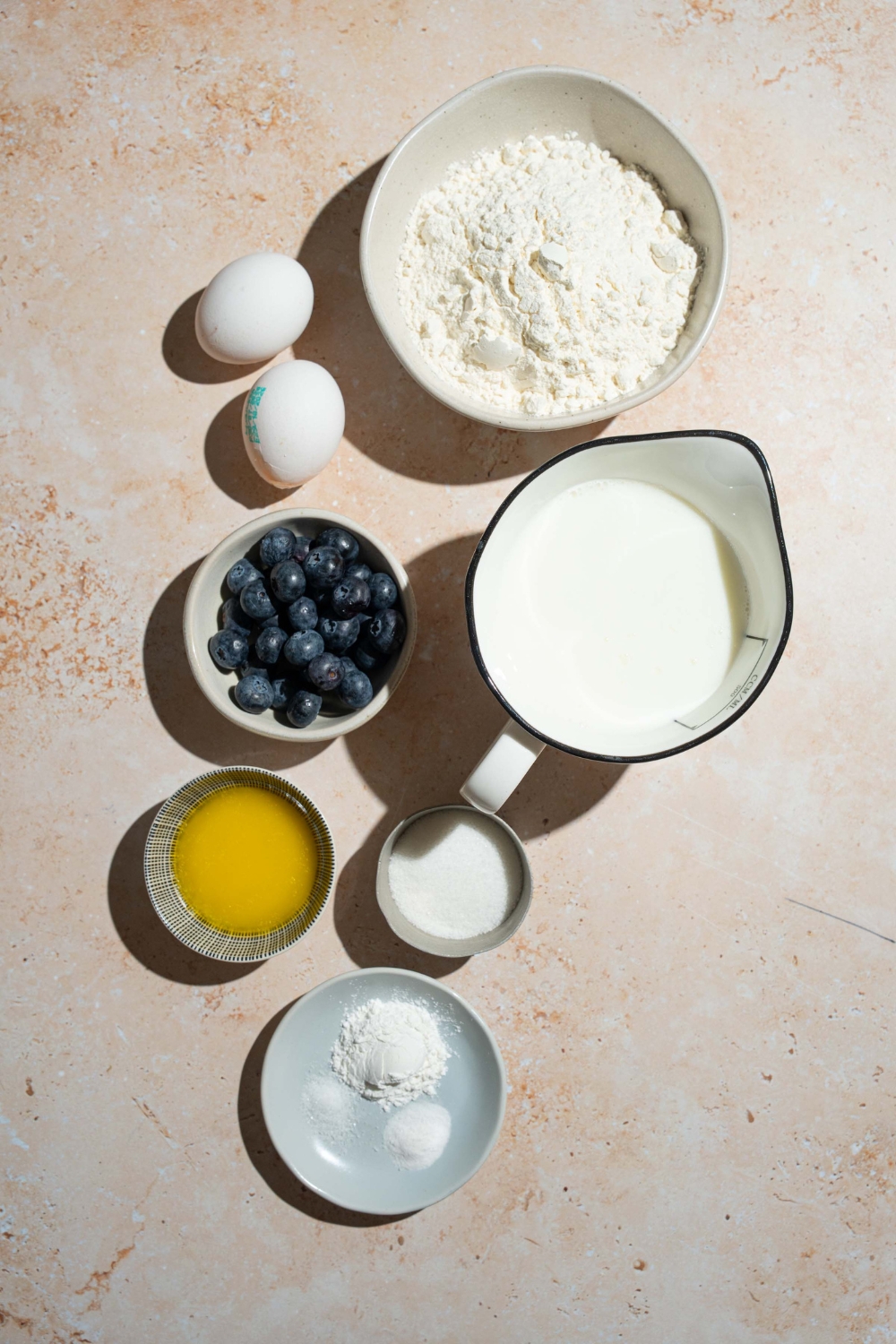 A tan counter with several bowls containing ingredients to make buttermilk blueberry pancakes including flour, buttermilk, eggs, blueberries, sugar, baking powder, baking soda, and salt.