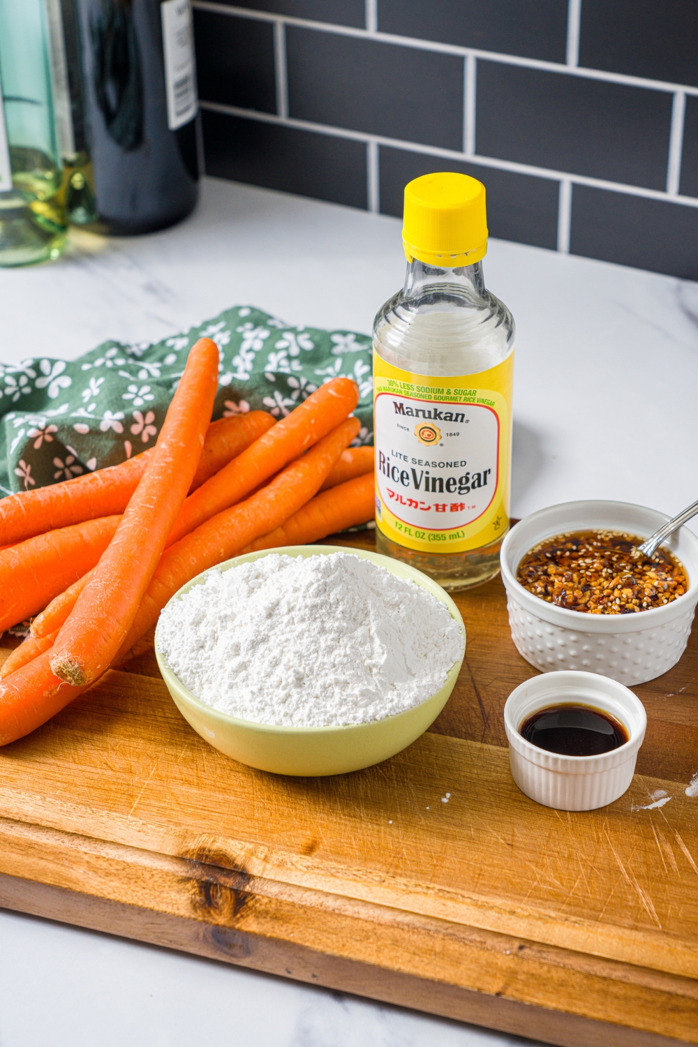 A wooden board with ingredients to make carrot button noodles including carrots, flour, chili crisp, soy sauce, and rice vinegar.