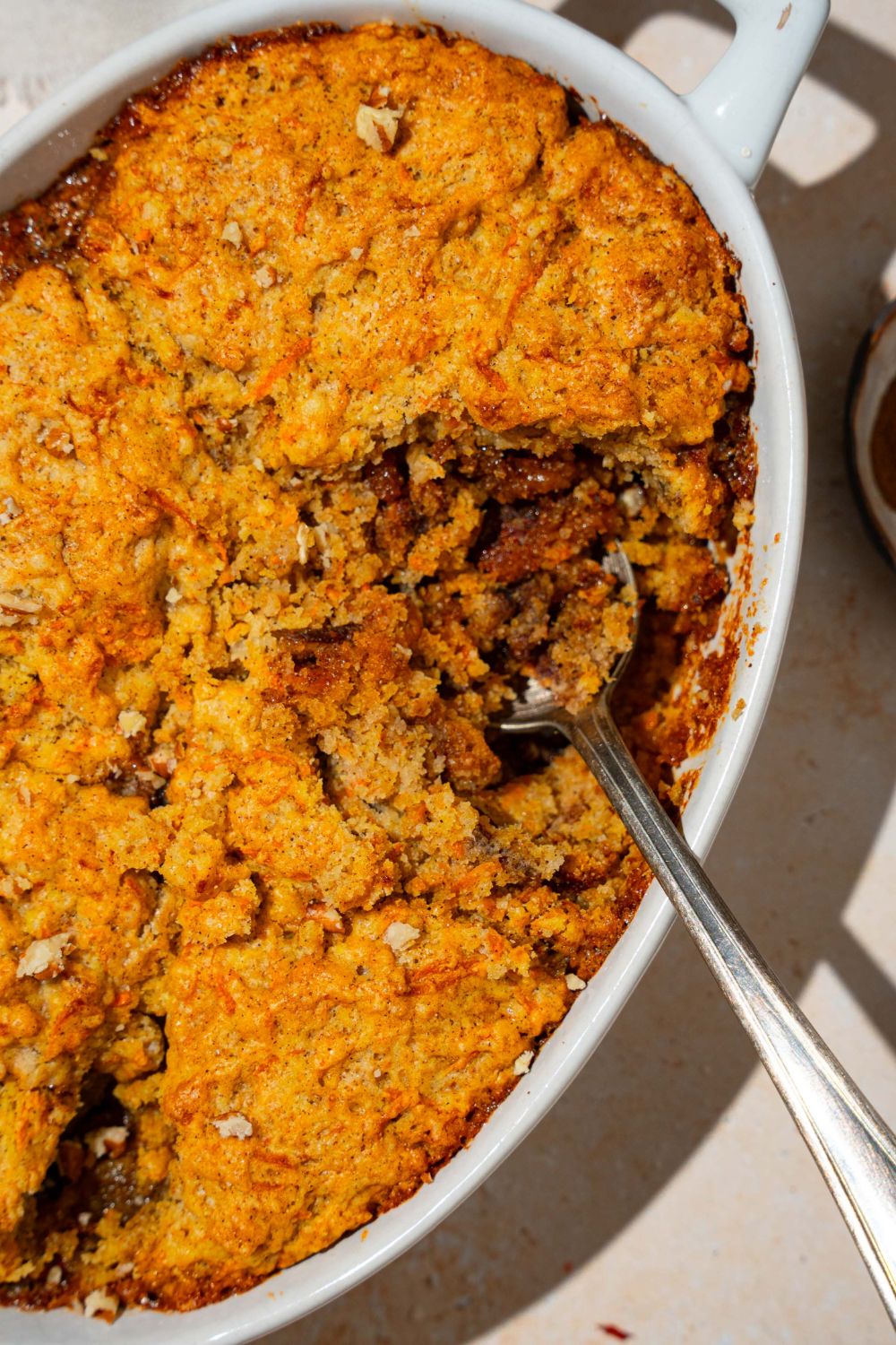 An oval baking dish with carrot cake cobbler with pecan pie filling. A spoon is dipped in the cobbler. The dish is on a tan counter.