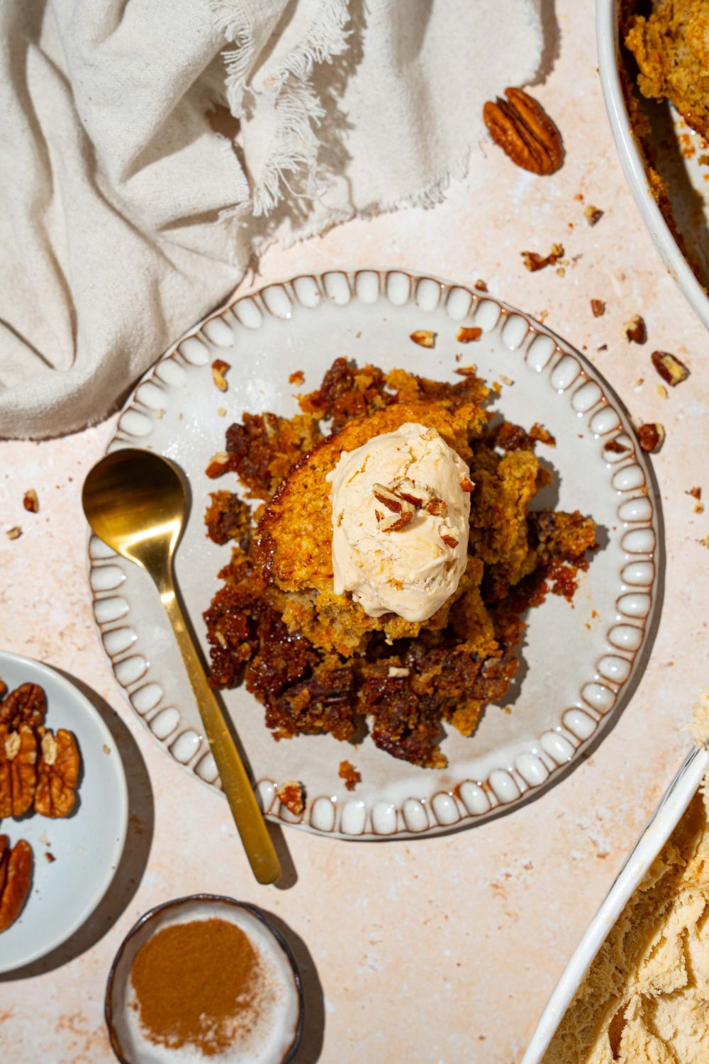 A white plate with a piece of carrot cake cobbler with a pecan filling topped with vanilla ice cream. There is a spoon on the plate. The plate is on a tan counter with a white cloth napkin.