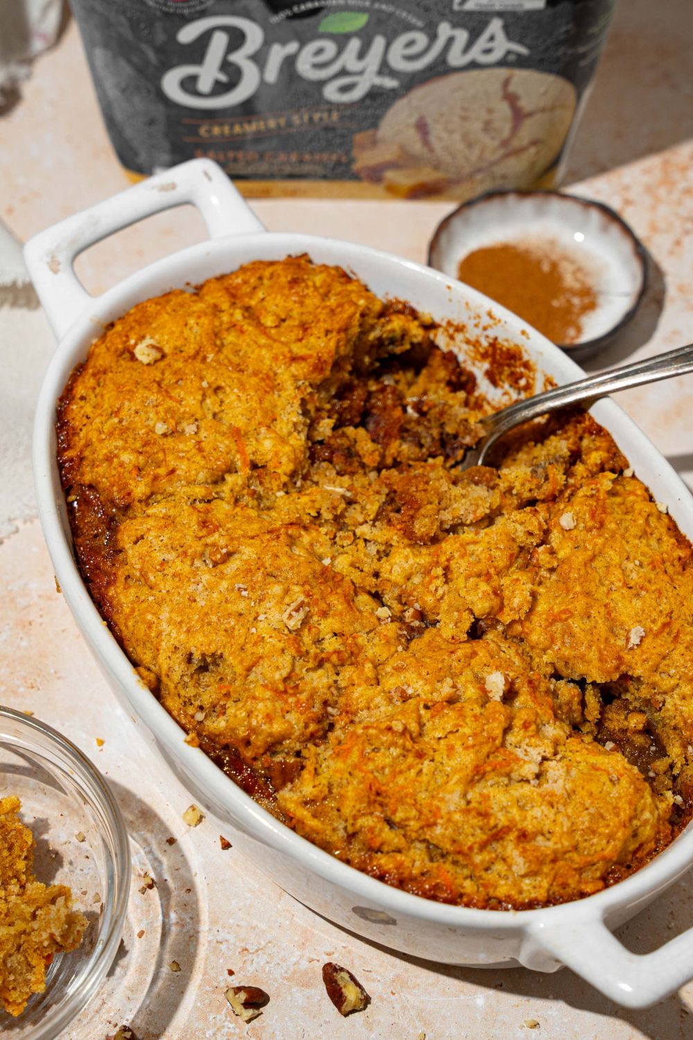 An oval baking dish with carrot cake cobbler with pecan pie filling. A spoon is dipped in the cobbler. The dish is on a tan counter with a white cloth napkin and bowl of pecans.