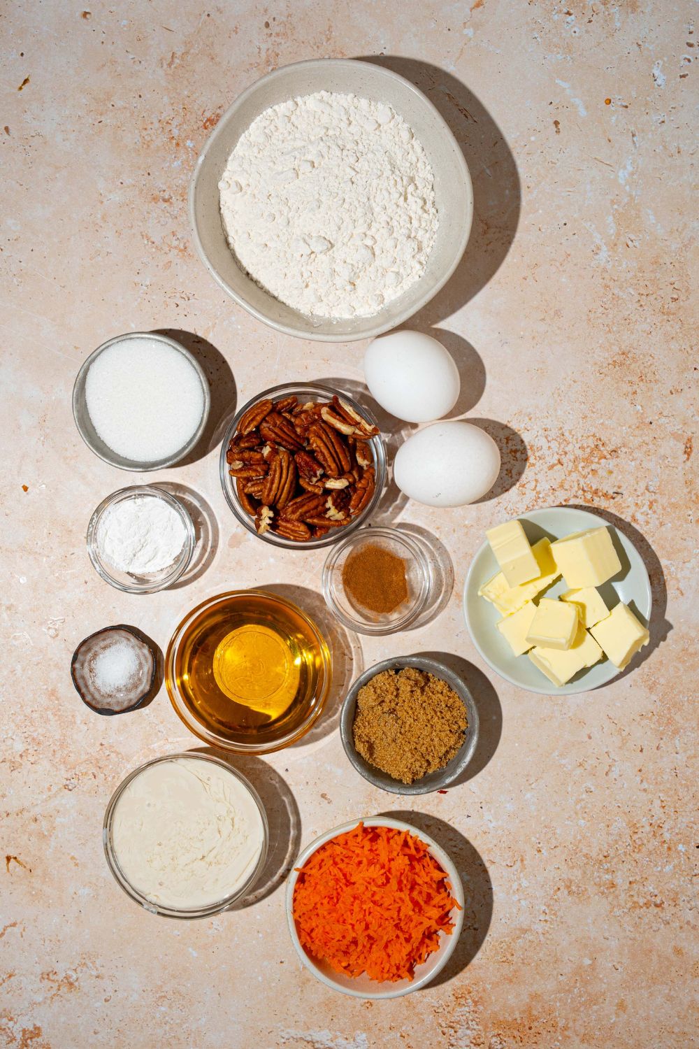 A tan counter with several bowls containing ingredients to make carrot cake cobbler with pecan pie filling including flour, eggs, brown sugar, vanilla extract, cinnamon, flour, butter, pecans, and spices.