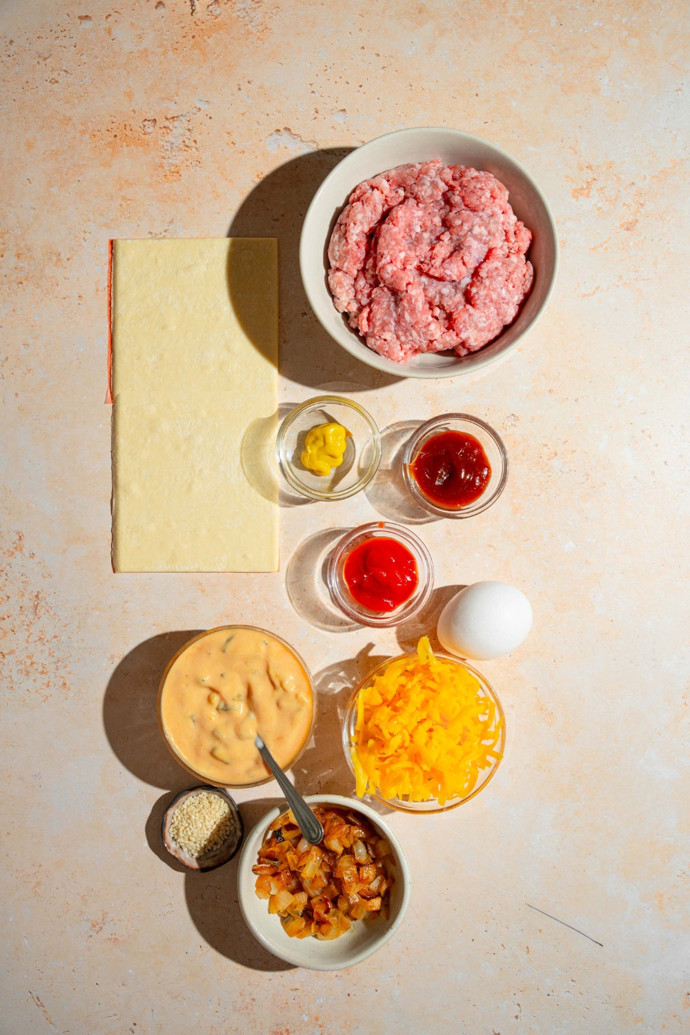 A tan counter with ingredients to make cheeseburger hot pockets including ground beef, puff pastry sheets, shredded cheese, caramelized onions, egg, and more.